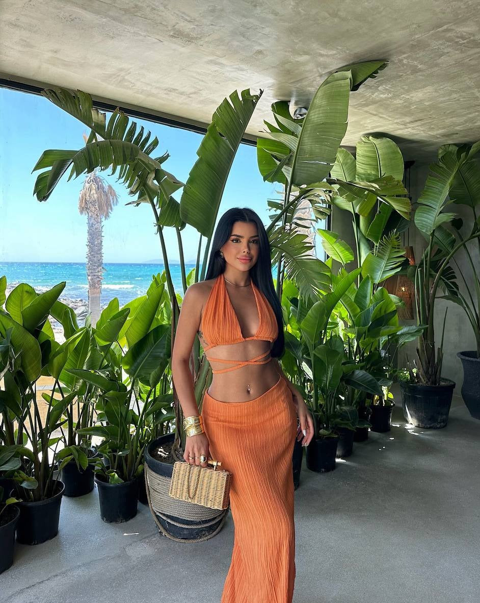 "Model wearing the Tropical Chic Set in orange, featuring a tie-up design and half-lined, zippered skirt, standing by a tropical resort."