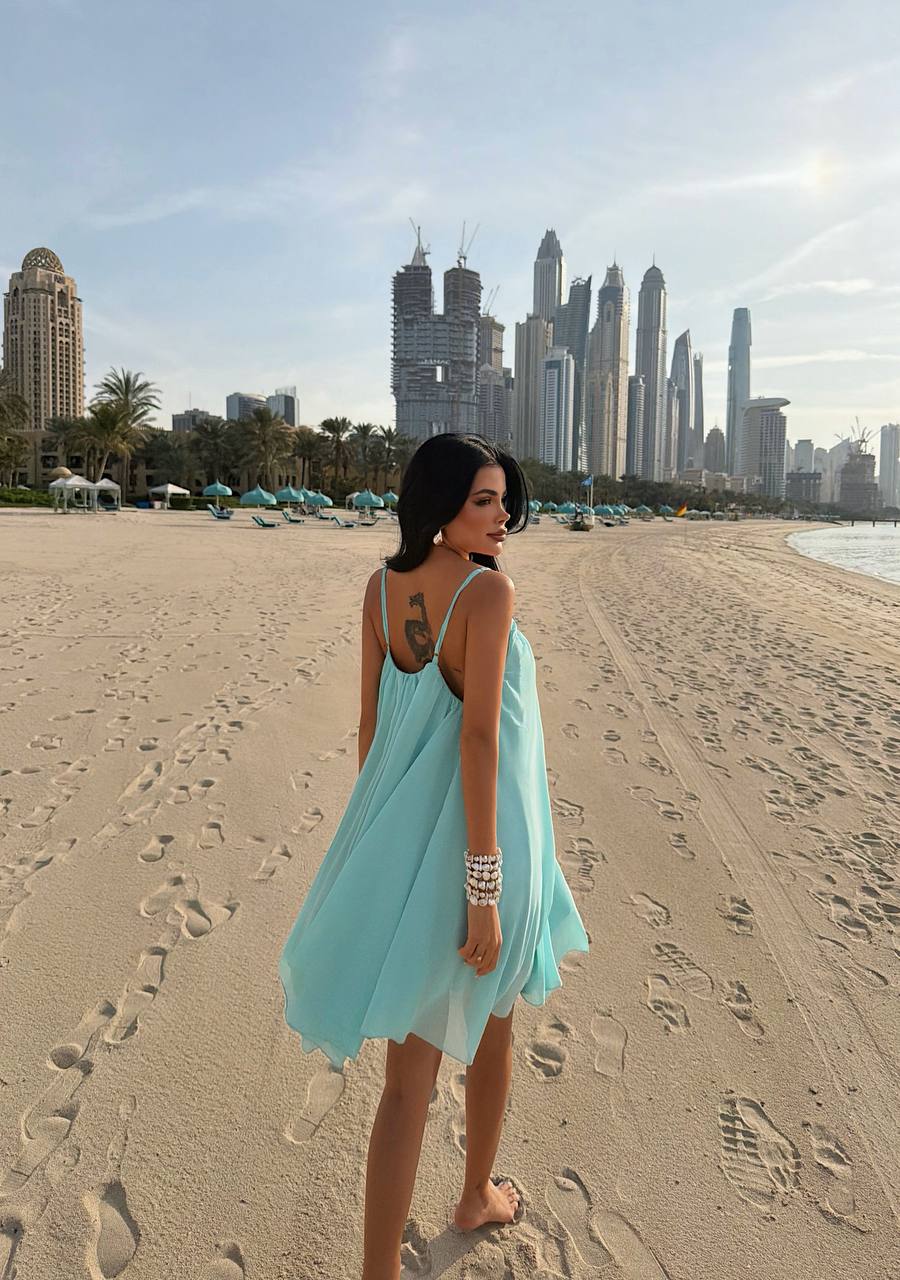 Woman in a light blue dress standing on a sandy beach with city skyline in the background