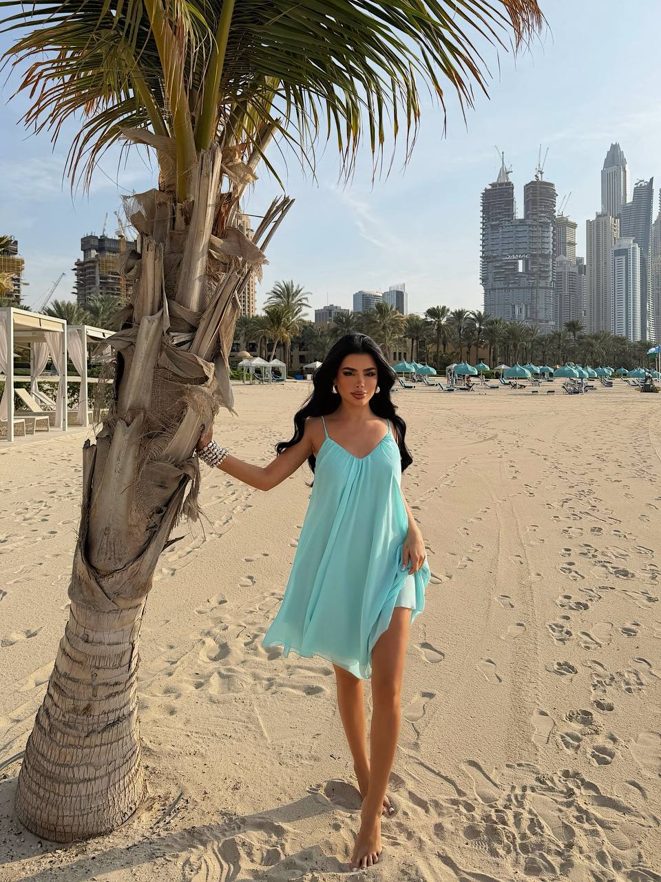 Woman in a light blue dress standing on a sandy beach with palm trees and city skyline in the background.