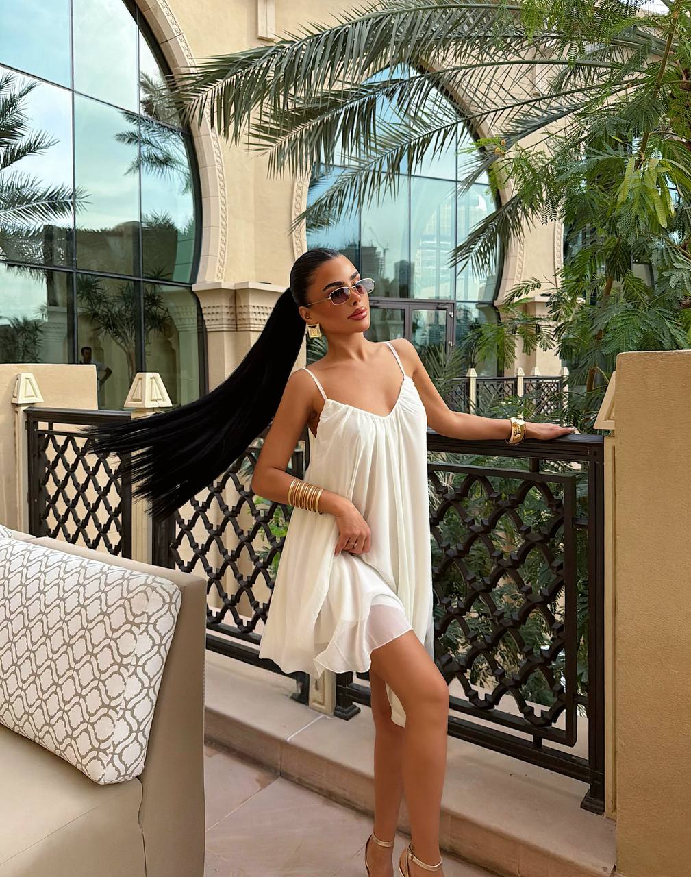 Woman in a white dress standing on a balcony with palm trees and a building in the background