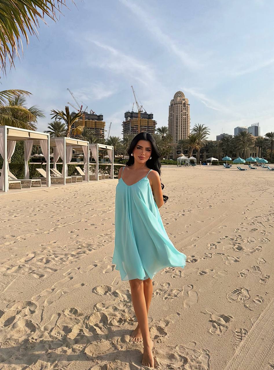 Woman in a light blue dress on a sandy beach with palm trees and buildings in the background