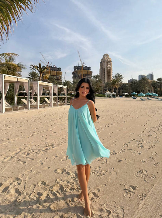 Woman in a light blue dress on a sandy beach with palm trees and buildings in the background