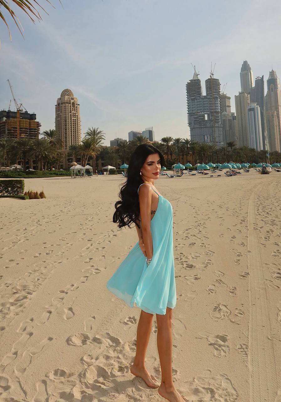 Woman in a light blue dress standing on a sandy beach with city skyline in the background